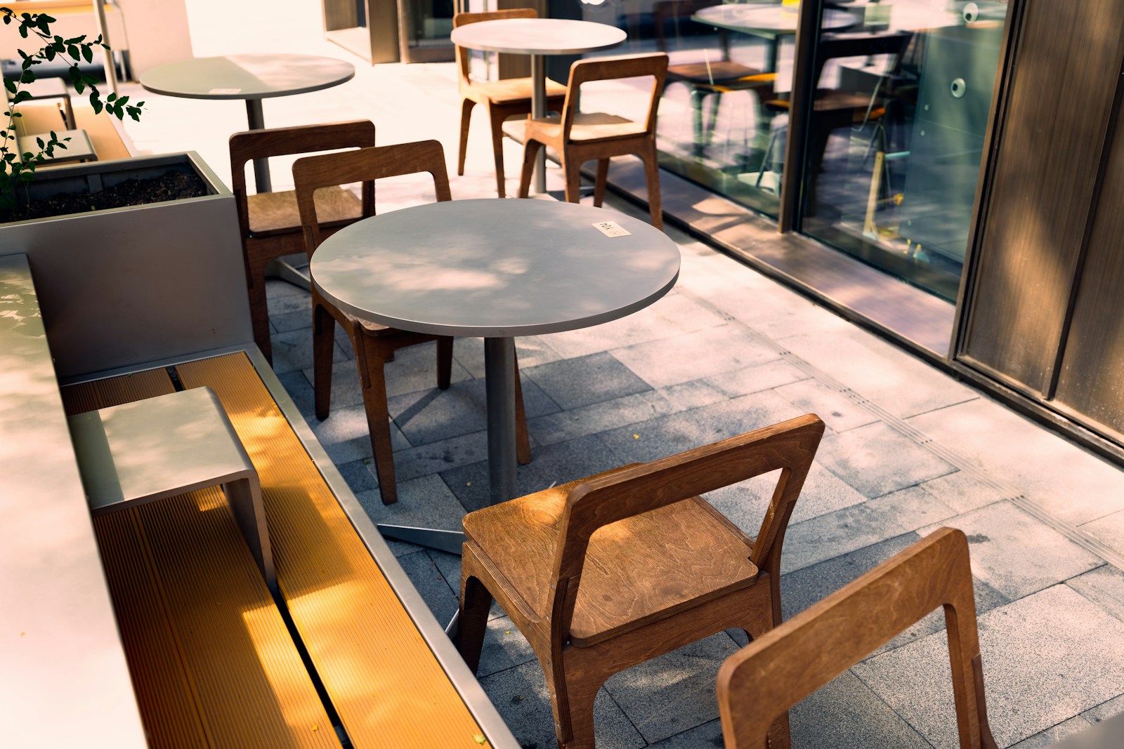 晴れた日の店先のテラス席。グレーの丸テーブルと木の椅子が並び、石畳に木漏れ日の影が落ちている