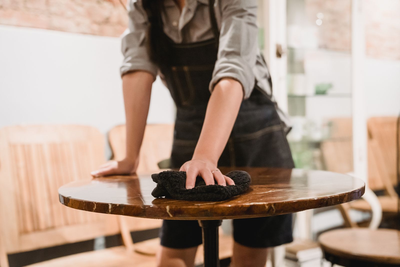 客のいないカフェで、エプロンをつけた店員が丸い木のテーブルを黒い布で拭いている手元